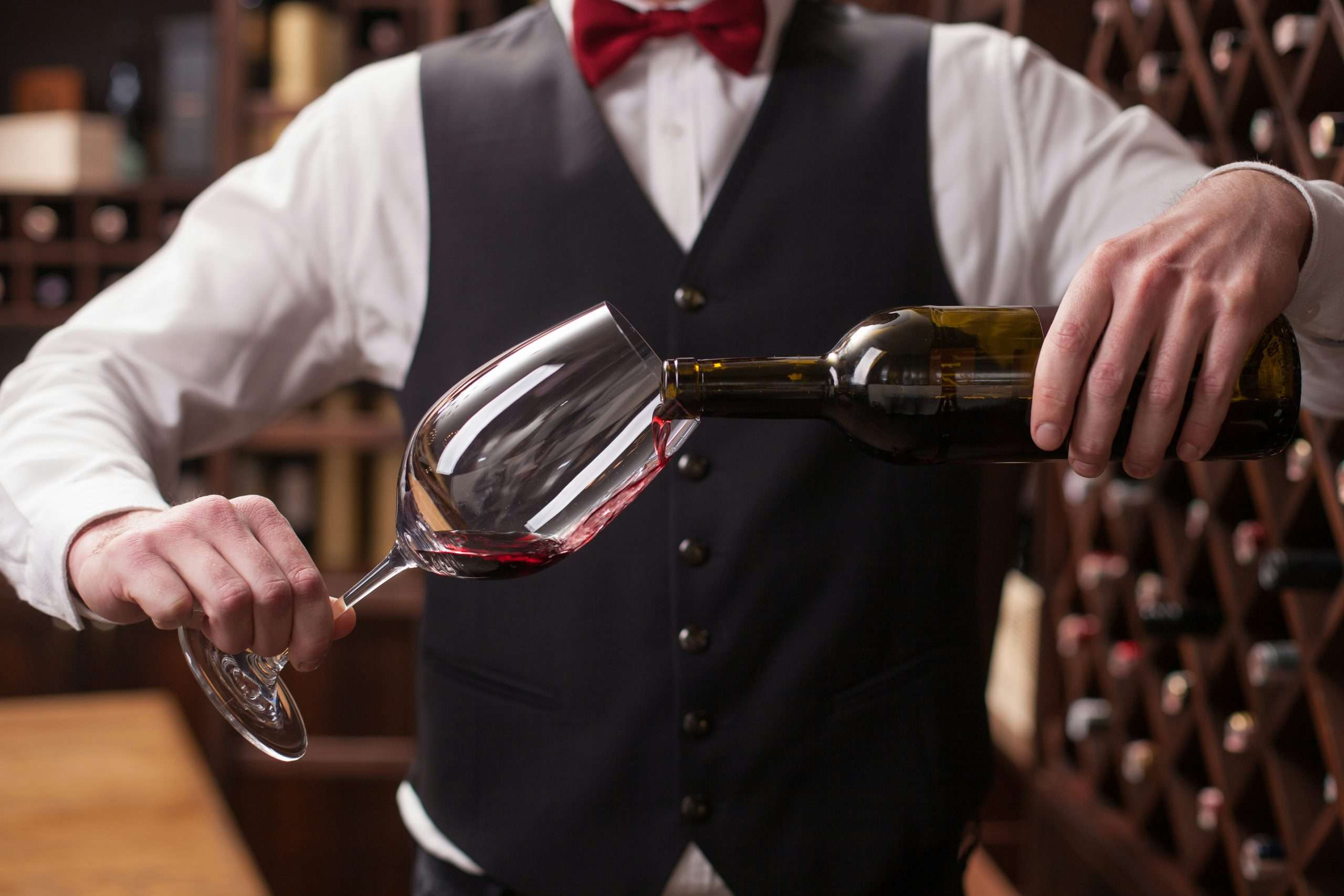A sommelier in a formal black vest, white shirt, and red bow tie pours red wine into a tilted glass with precision. The background features a wine cellar with wooden racks filled with bottles, creating an elegant and refined atmosphere.
