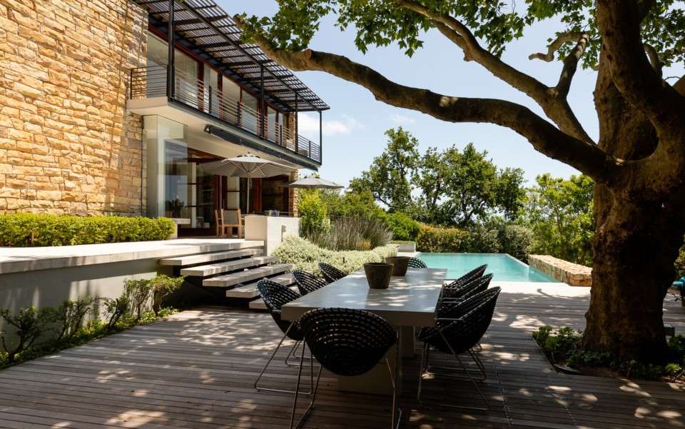 Shaded outdoor dining area under a large tree at a luxury Cape Town villa with pool and garden views.