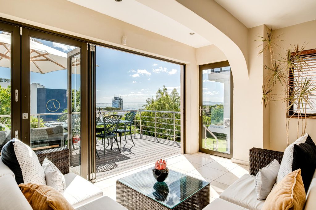 Kaya Eden – Bright indoor lounge opening through folding glass doors onto a sunny balcony with elevated views towards the Atlantic Ocean.