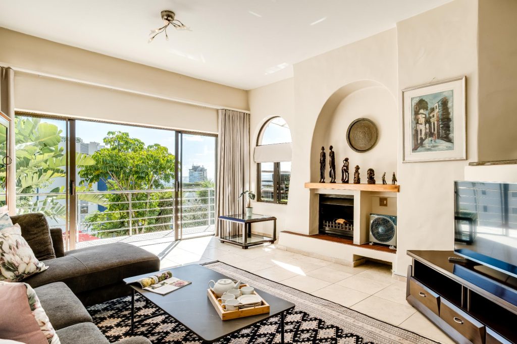 Kaya Eden – Comfortable living room with fireplace, large television and sliding glass doors opening onto a balcony with leafy neighbourhood and ocean views.