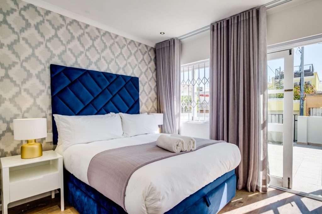 De Waterkant DW-001 – Main Bedroom – Elegant double bedroom with a striking upholstered headboard, soft neutral finishes and doors opening onto a private outdoor area.