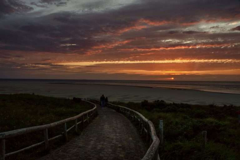 Couple walking on a promenade at sunset with a scenic beach and vibrant sky.