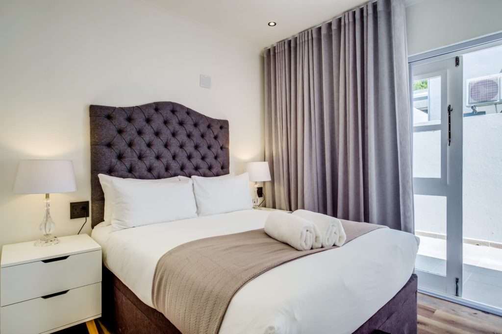 De Waterkant DW-001 – Double Bedroom – Comfortable double bedroom with a plush upholstered headboard, contemporary décor and doors opening outside.