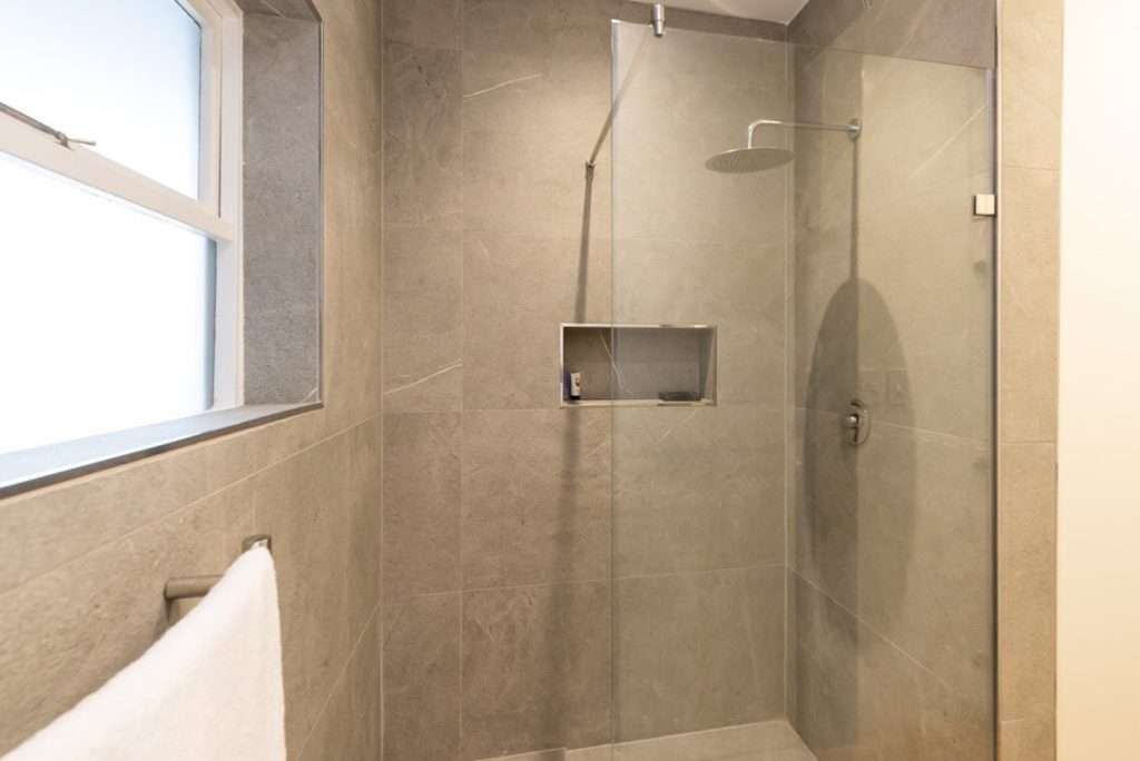Mimosa Views – Contemporary bathroom featuring elegant grey stone-effect tiling, a spacious glass-enclosed walk-in shower, rainfall showerhead and natural light.