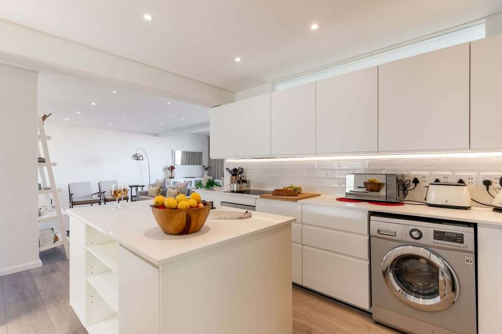 Protea Views Apartment – Modern open-plan kitchen with sleek white cabinetry, a central island, contemporary appliances and a washing machine.