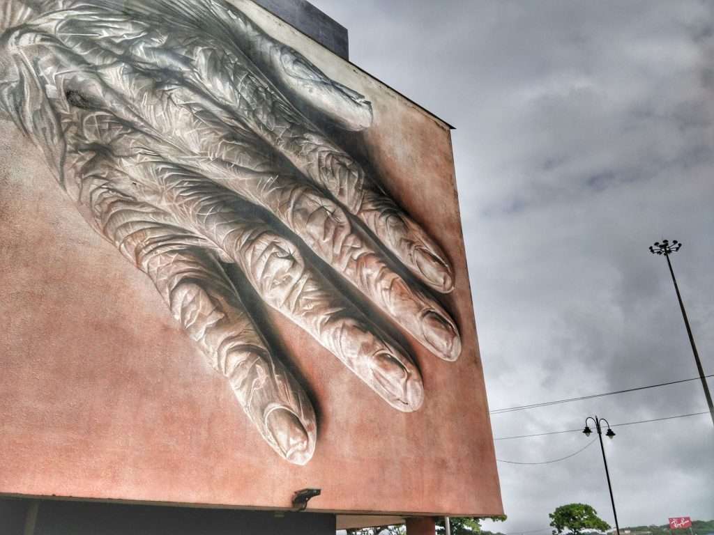 A massive mural of a hand on an urban building exterior beneath a cloudy sky.