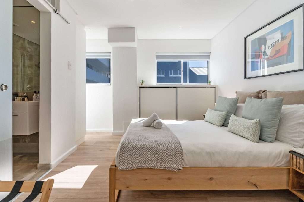 Protea Views Apartment – Tranquil bedroom with a wooden bed frame, soft neutral furnishings, natural light and an adjoining en-suite bathroom.