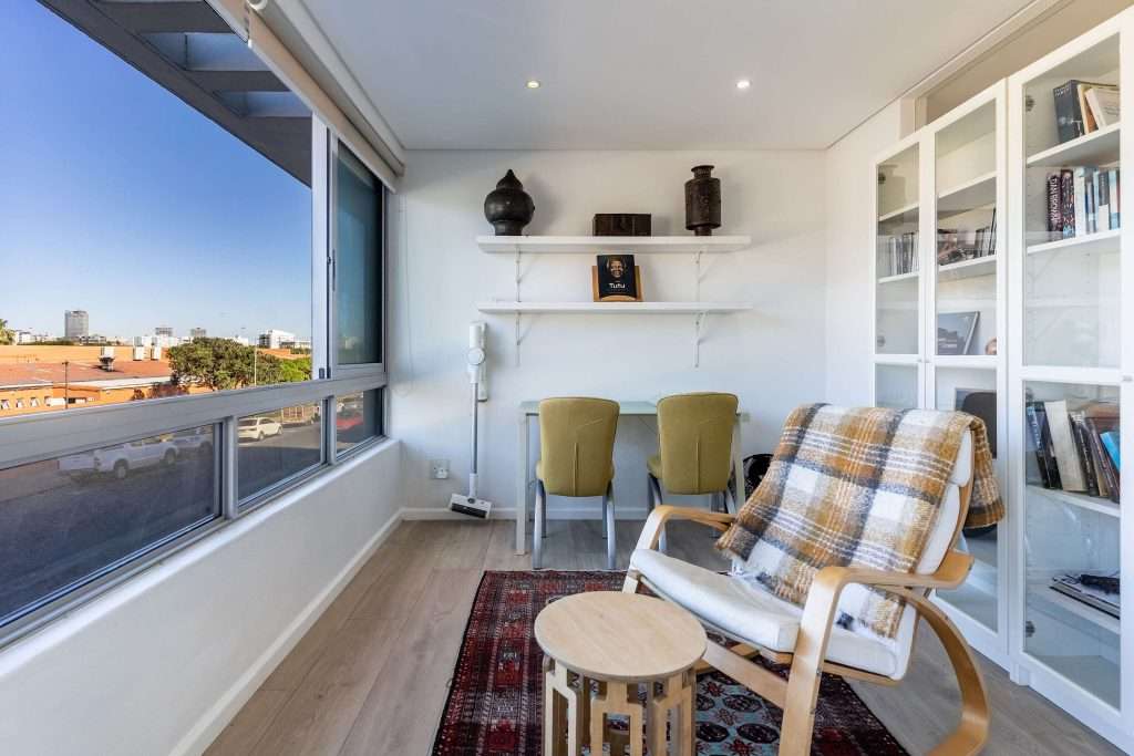 Protea Views Apartment – Cosy enclosed sunroom with comfortable seating, bookshelves, a small desk and neighbourhood views, ideal for reading or relaxing.